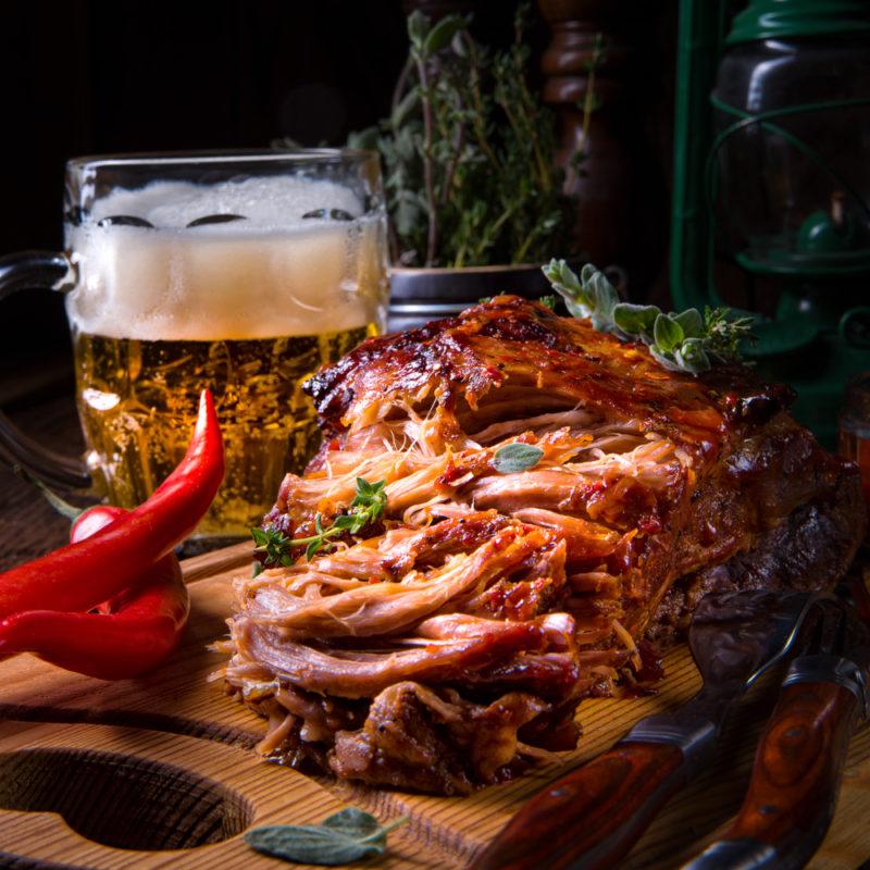 Saftiges Pulled Pork Fleisch auf einer Holzplatte neben einem großen goldgelben Bier